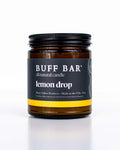 Buff Bar All-natural candle in a amber glass with black lid and label featuring the Lemon Drop fragrance and the Buff Bar Bison illustration. The candle is on a white background.