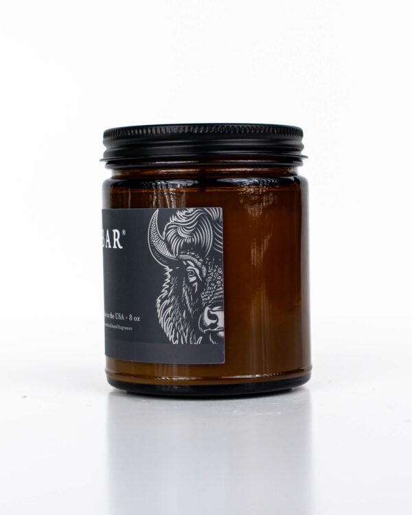 Buff Bar All-natural candle in a amber glass with black lid and label featuring the Trail Ride fragrance and the Buff Bar Bison illustration. The candle is on a white background.
